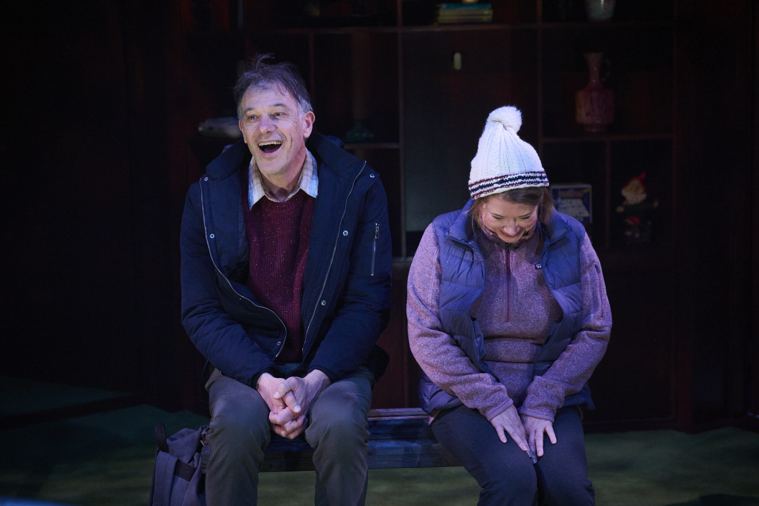 A man and woman sit on a bench. The man, wearing a jacket, smiles and laughs, whilst the woman, in a knitted hat and waistcoat, looks down and smiles shyly. They appear to be sharing a light-hearted moment together.