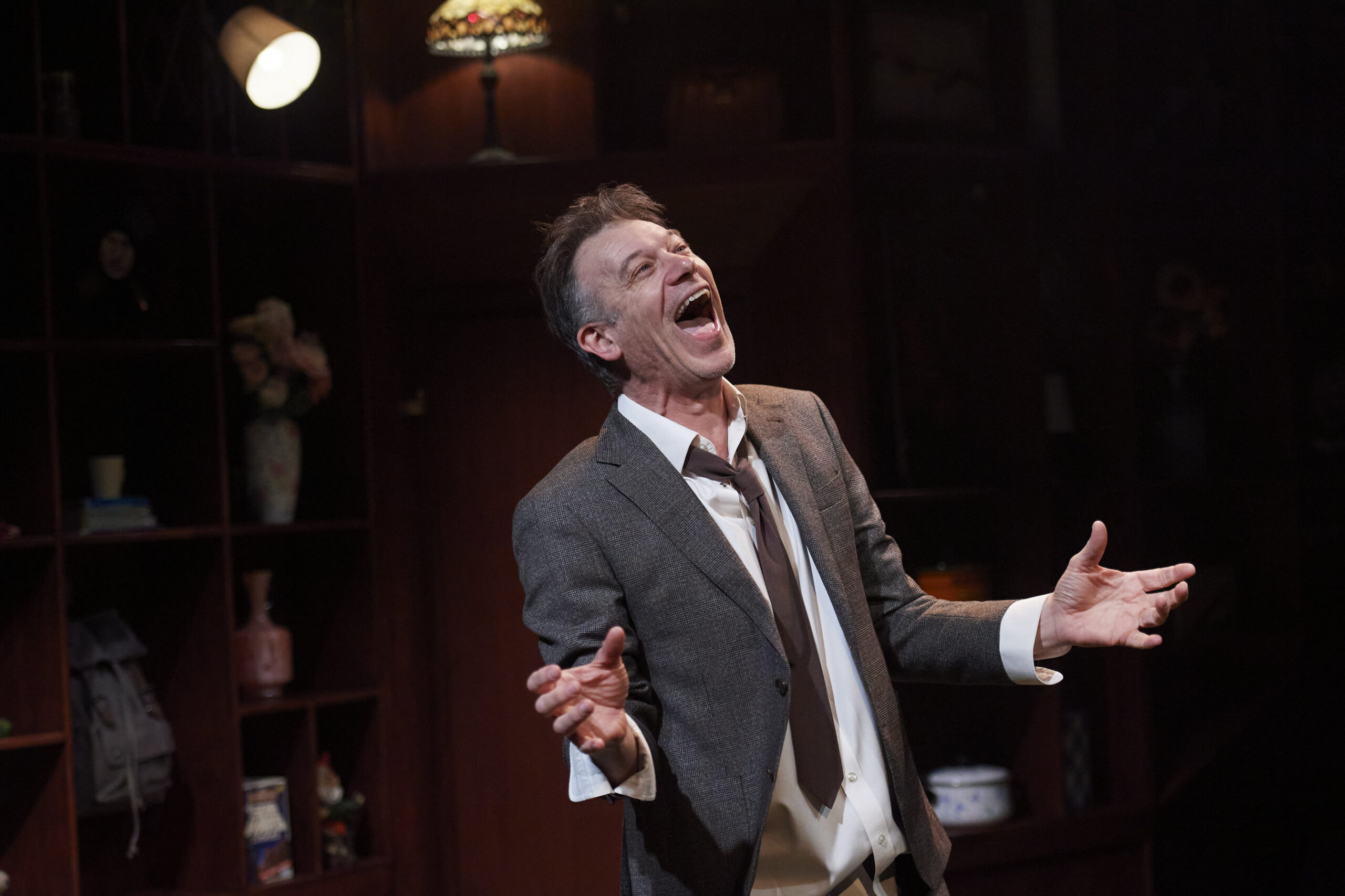A man in a grey suit and loosened tie stands indoors with his mouth open wide and eyes closed, appearing to sing or shout passionately. He gestures with both hands. Shelves and dim lighting are visible in the background.