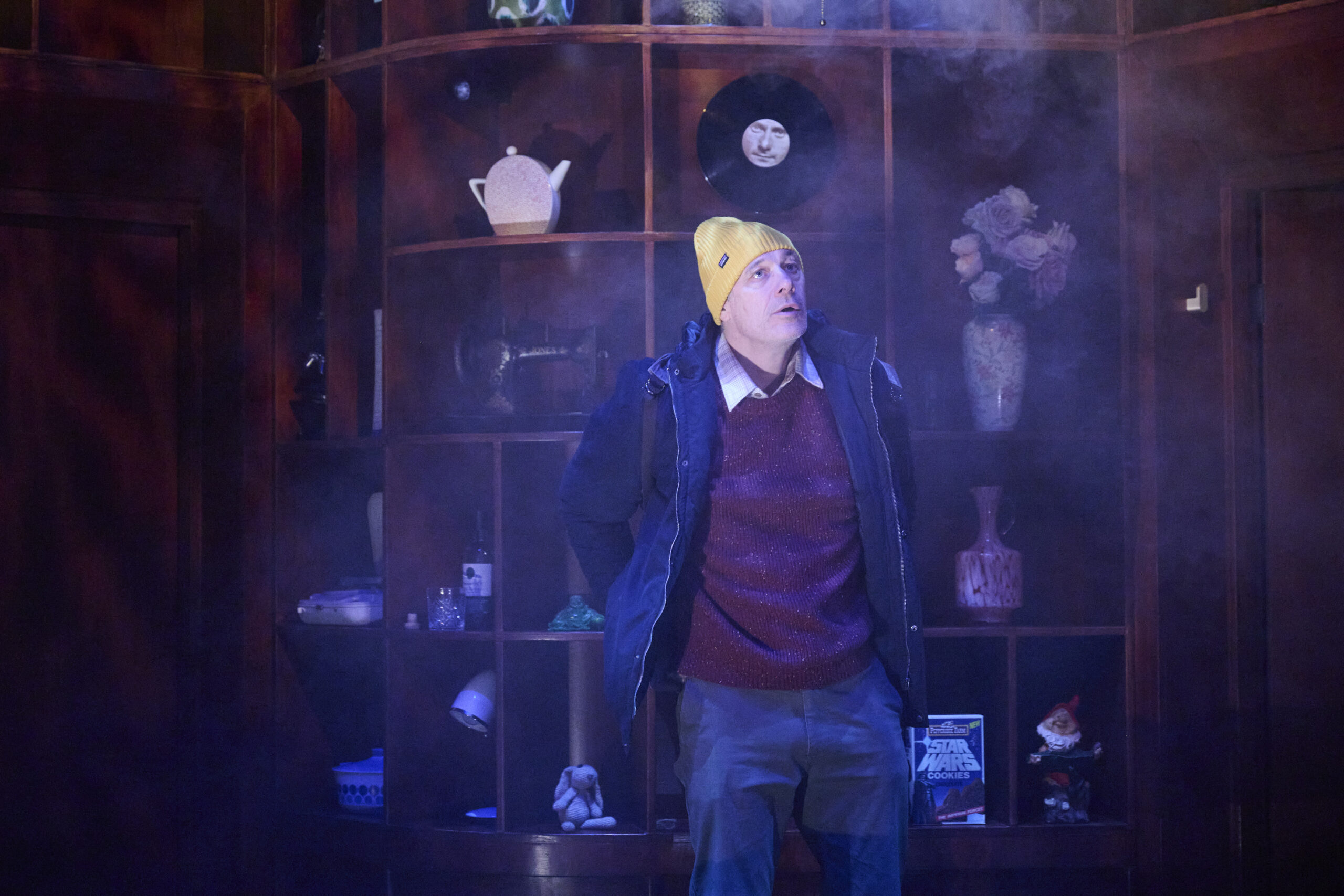 A person wearing a yellow beanie, maroon jumper, and blue jacket stands in front of a wooden shelf filled with various objects, including vases, a teapot, and figurines, surrounded by a misty atmosphere.