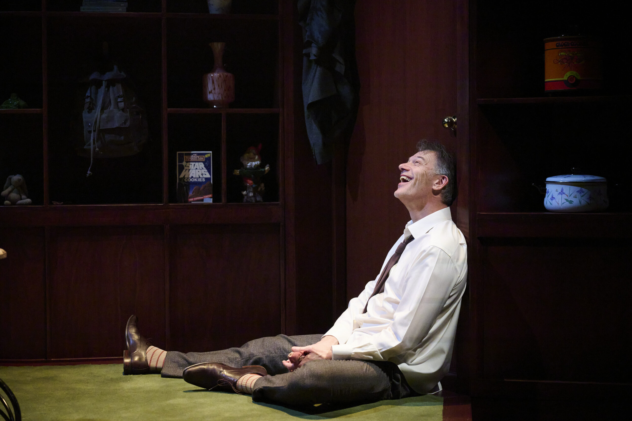 A man in a white shirt and tie sits on the floor, leaning against a wooden cupboard, laughing. Behind him are shelves with various objects, including books, a jar, and a rucksack.