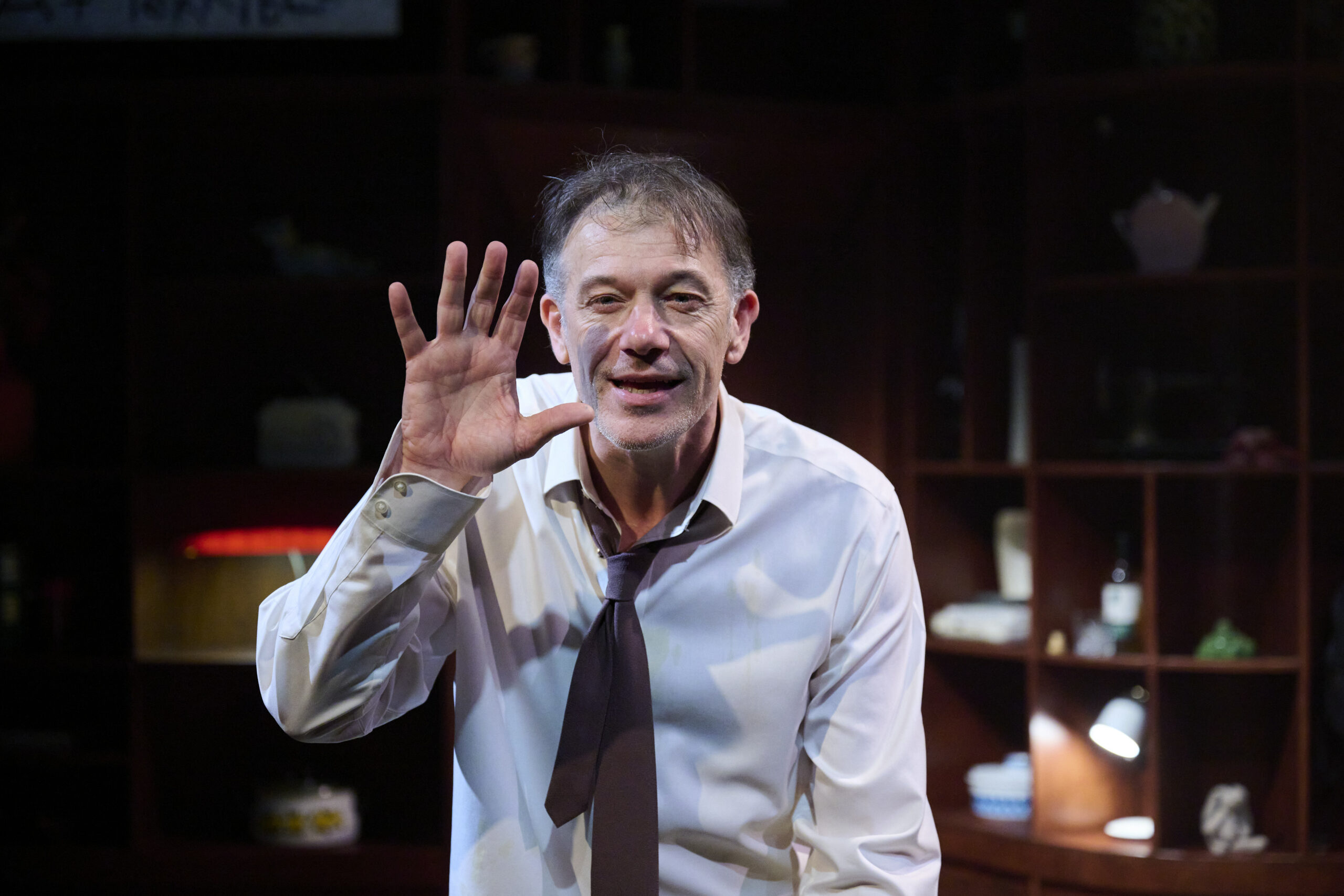A man in a white shirt and loose dark tie stands indoors, holding his hand up with fingers spread, as if waving or signalling. He is smiling slightly, and shelves with various objects are visible in the dim background.