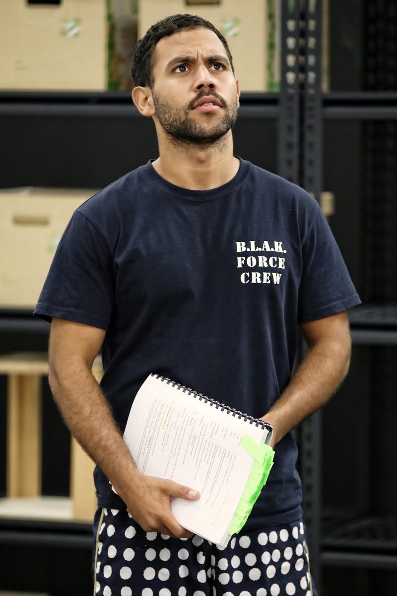 A man with short hair and a beard, wearing a navy B.L.A.K. FORCE CREW t-shirt and polka-dot shorts, stands indoors holding a spiral-bound document with green tabs. Shelves and cardboard boxes are in the background.