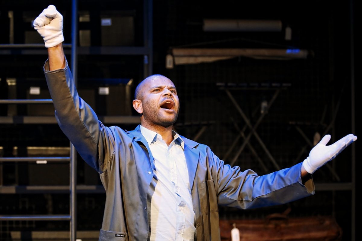 A man wearing a grey coat and white gloves raises one fist and gestures with his other hand whilst singing or speaking passionately on stage. A dark shelving unit is visible in the background.