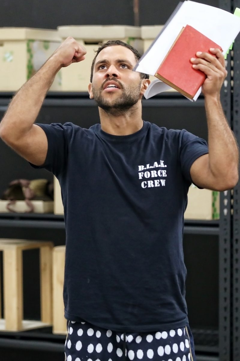 A man wearing a B.L.A.K. FORCE CREW T-shirt and polka-dot shorts raises a fist and holds two books, looking upwards with a passionate expression. Shelves and boxes are visible in the background.