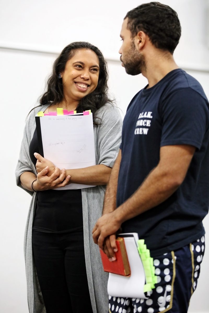Two people stand and talk, holding notebooks with colourful sticky notes. The woman smiles whilst the man listens. They appear to be in a casual, indoor setting, possibly collaborating or discussing something together.