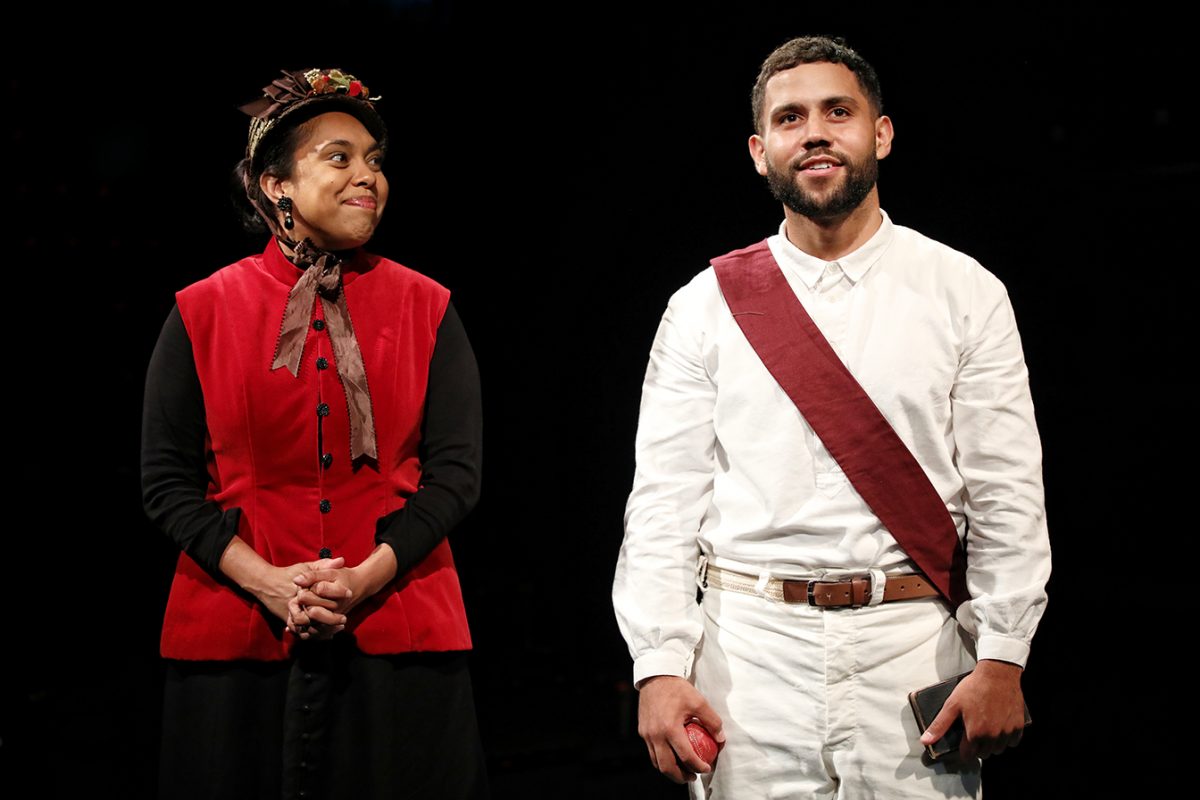 Two actors on stage, one wearing a red waistcoat, brown hat, and brown necktie, smiling at the other, who wears a white outfit with a maroon sash and holds a small book and red ball, both against a dark background.