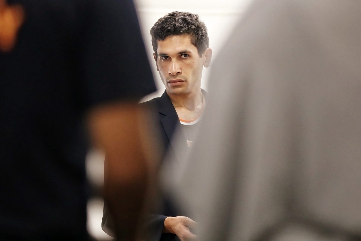 A man with short curly hair and a serious expression looks forward, partially obscured by two blurred figures in the foreground. The background is plain and softly lit.