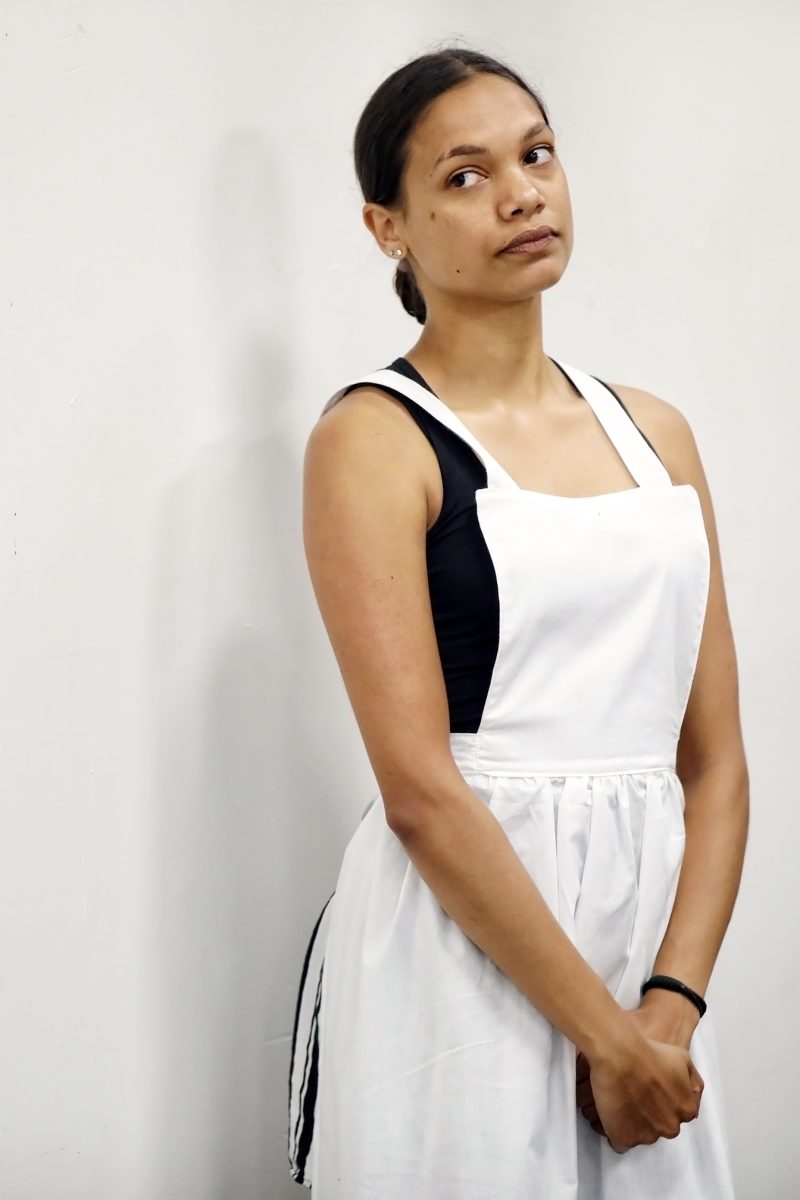 A woman with medium skin and dark hair tied back stands against a plain white wall, wearing a white apron over a black top. She looks slightly upwards with a neutral or contemplative expression, hands folded in front.