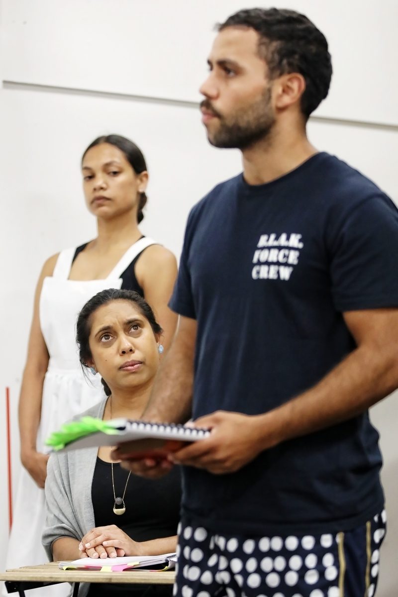Three people are indoors. A man in the foreground, holding papers, speaks while two women in the background listen; one stands in a white dress, and the other sits with a notebook, looking up at him.