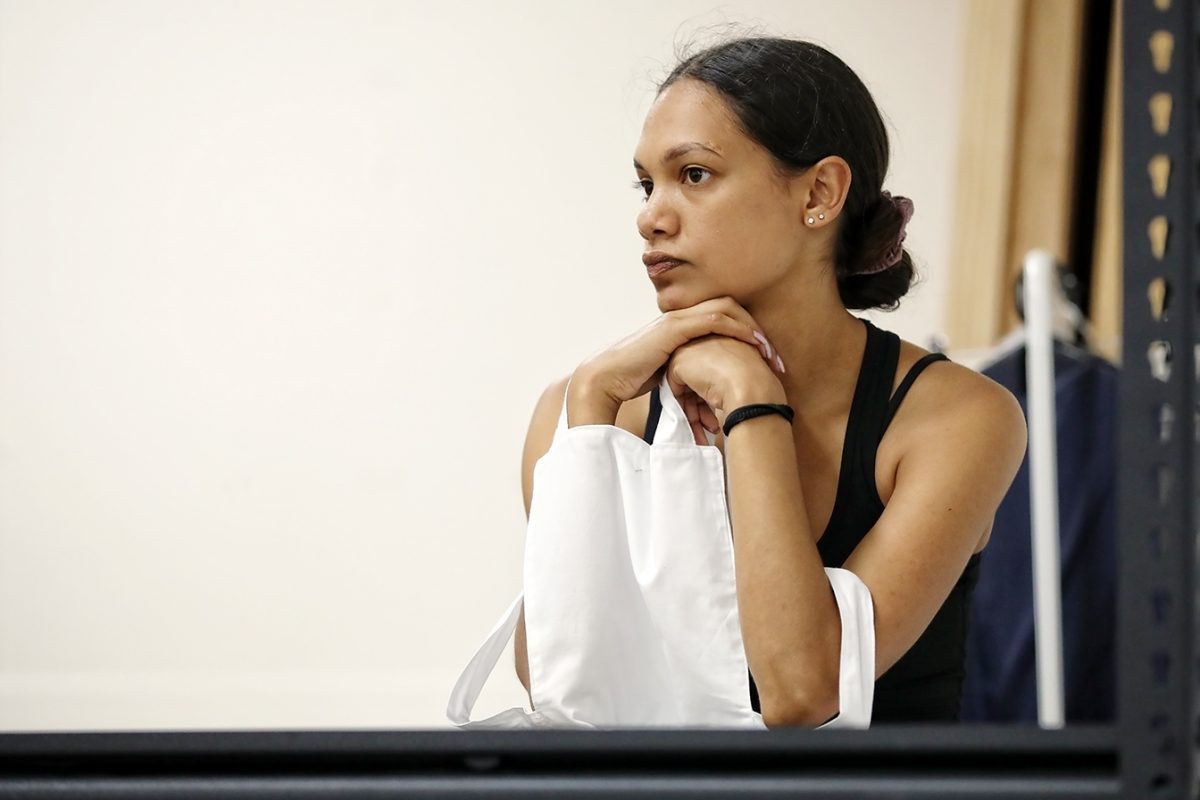 A woman with her arm in a white sling rests her chin on her hands, looking thoughtfully to the side. She has dark hair pulled back and wears a black vest top. The background is neutral and uncluttered.