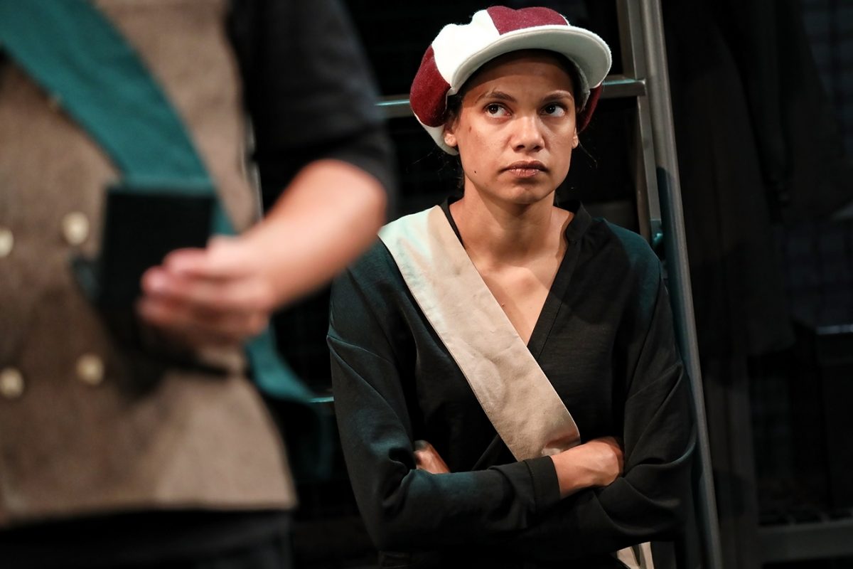 A person wearing a two-toned cap and a sash over a dark shirt stands with arms folded, looking serious. Another person, partially visible, is in the foreground holding an object.