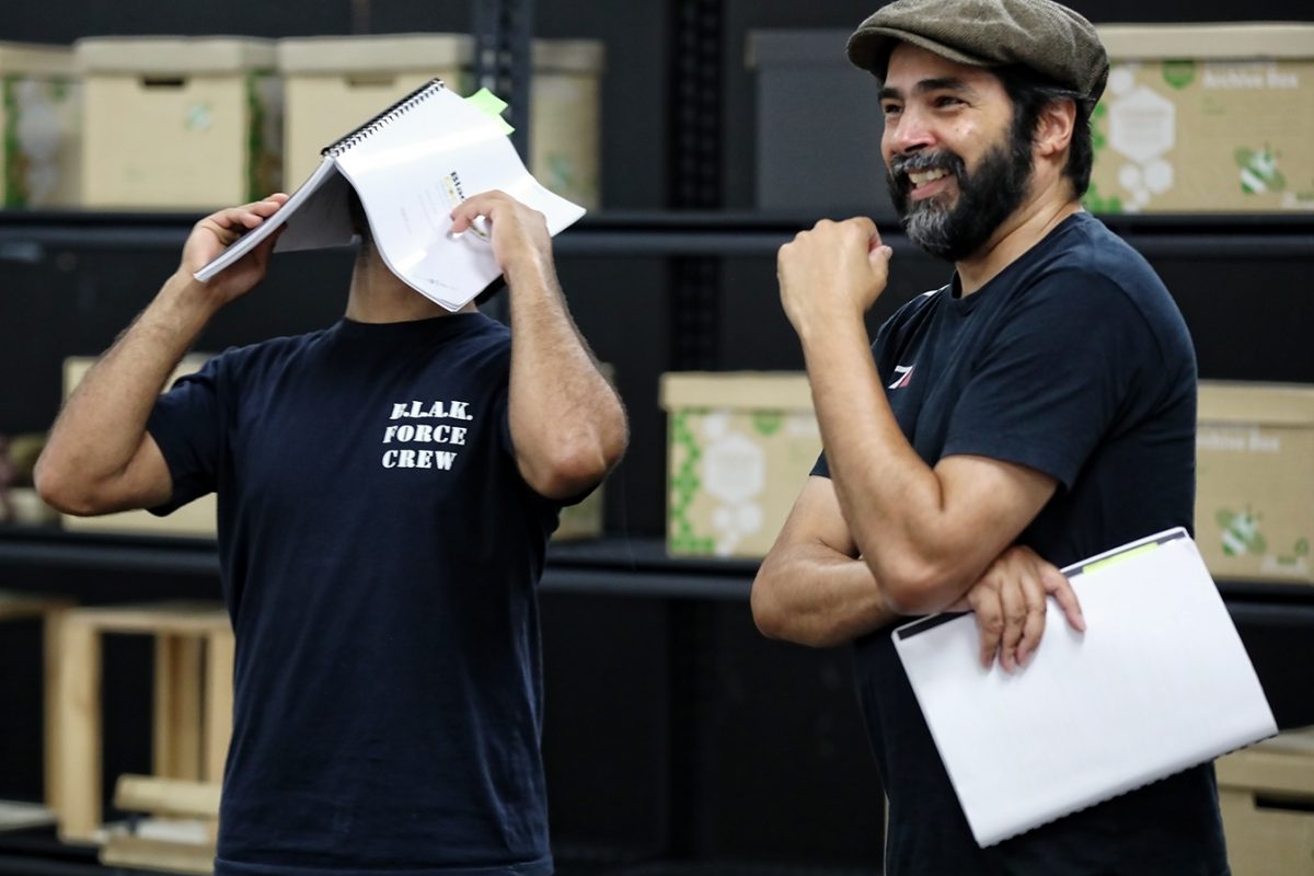 Two men stand indoors; one covers his face with a script, wearing a BLACK FORCE CREW t-shirt. The other, holding papers and wearing a cap, smiles. Shelves with storage boxes are in the background.