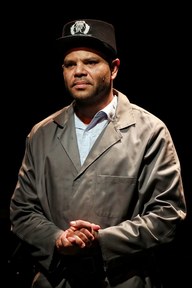 A man wearing a dark cap and a grey buttoned jacket stands with his hands clasped, looking thoughtful under a spotlight against a dark background.