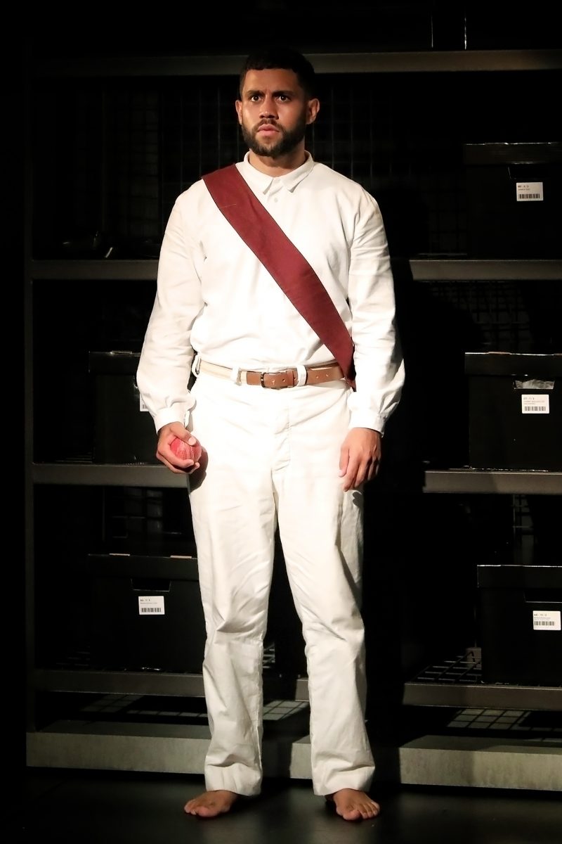 A man stands barefoot on stage wearing a white outfit with a brown diagonal sash and holding a red apple. Behind him are shelves filled with black storage boxes. He has a serious expression and is lit by stage lighting.