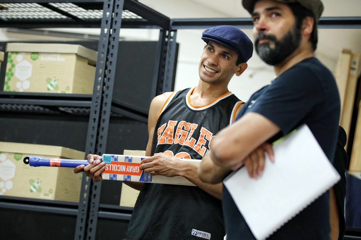 Two men stand together indoors; one wears a sports kit and cap, smiling while holding a cricket bat. The other, bearded and wearing a dark shirt and cap, holds a notebook. Shelves and boxes are visible in the background.