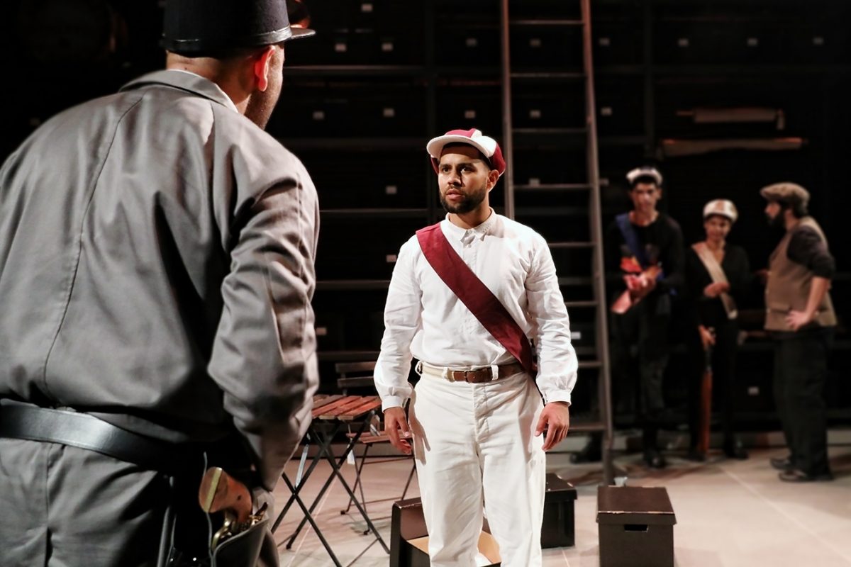 A man in a white uniform and cap with a red sash stands facing another man in grey, while three blurred figures watch in the background on a dimly lit stage.
