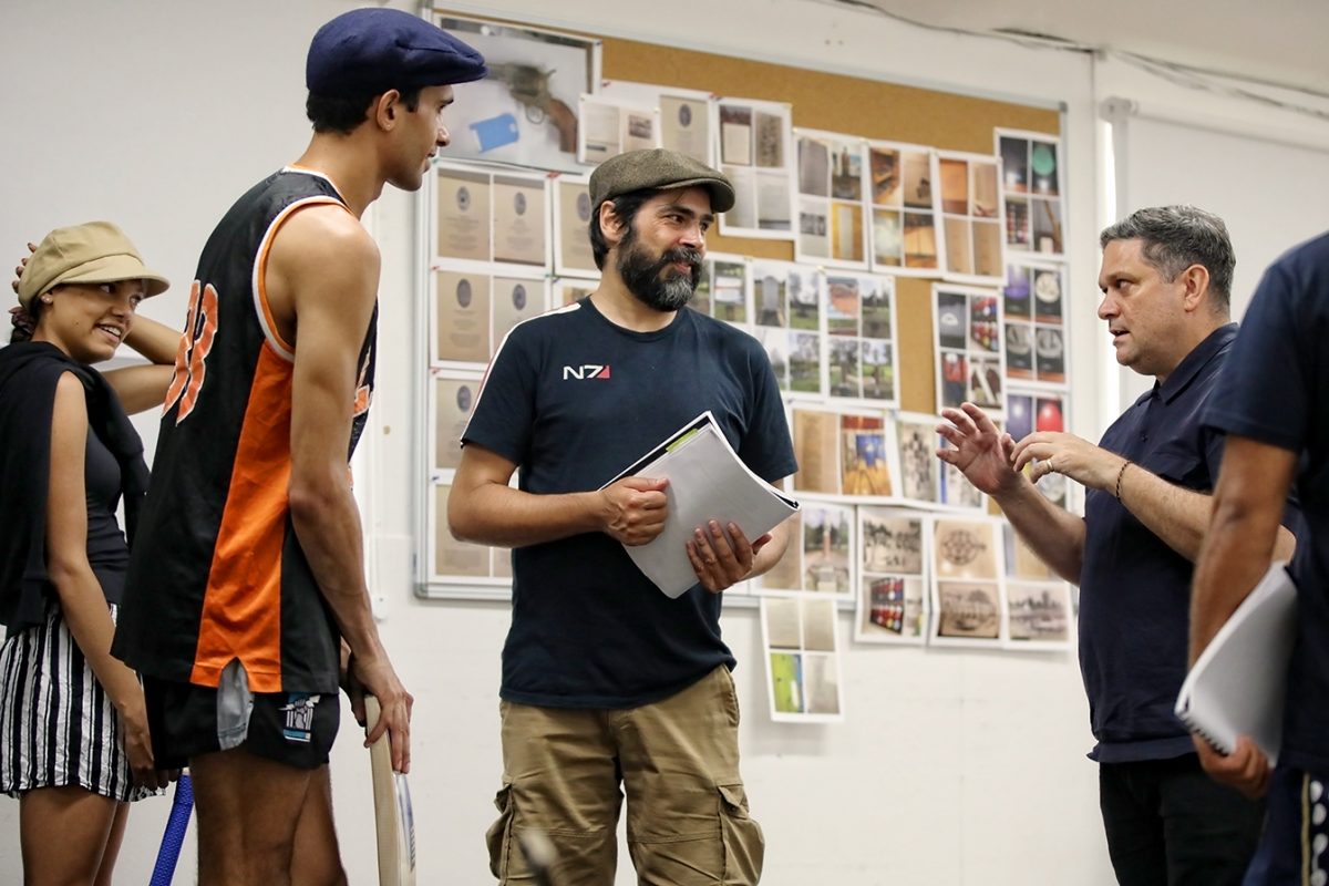 A group of four people are talking in a room with papers and photos pinned to a wall. Two people hold notebooks, and one wears a sports shirt and holds crutches. They appear to be in a discussion or workshop.