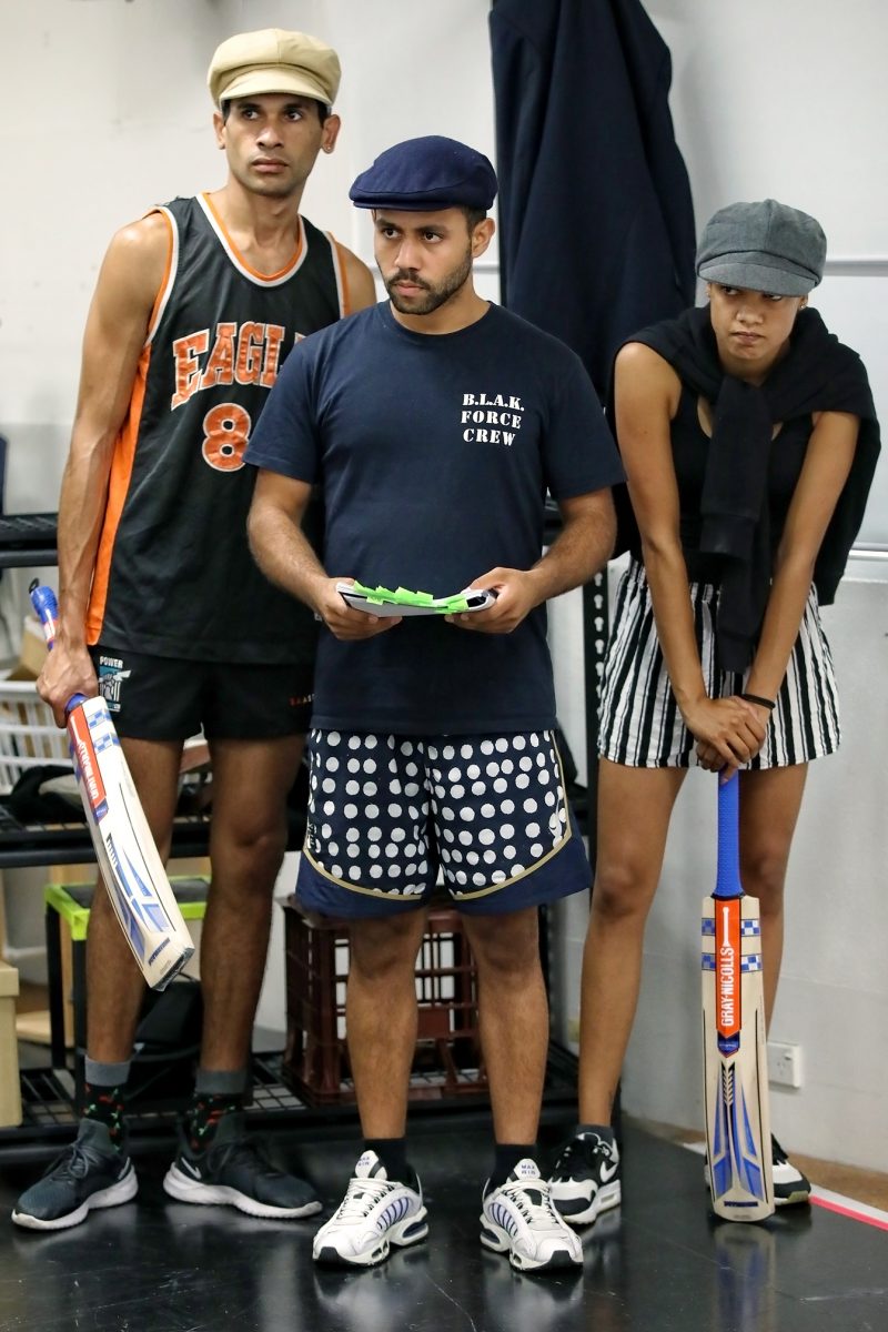 Three people stand indoors holding cricket bats. Two wear shorts, vests, and flat caps; one wears striped shorts, a jumper over her shoulders, and a cap. They look attentive, standing close together in a casual setting.