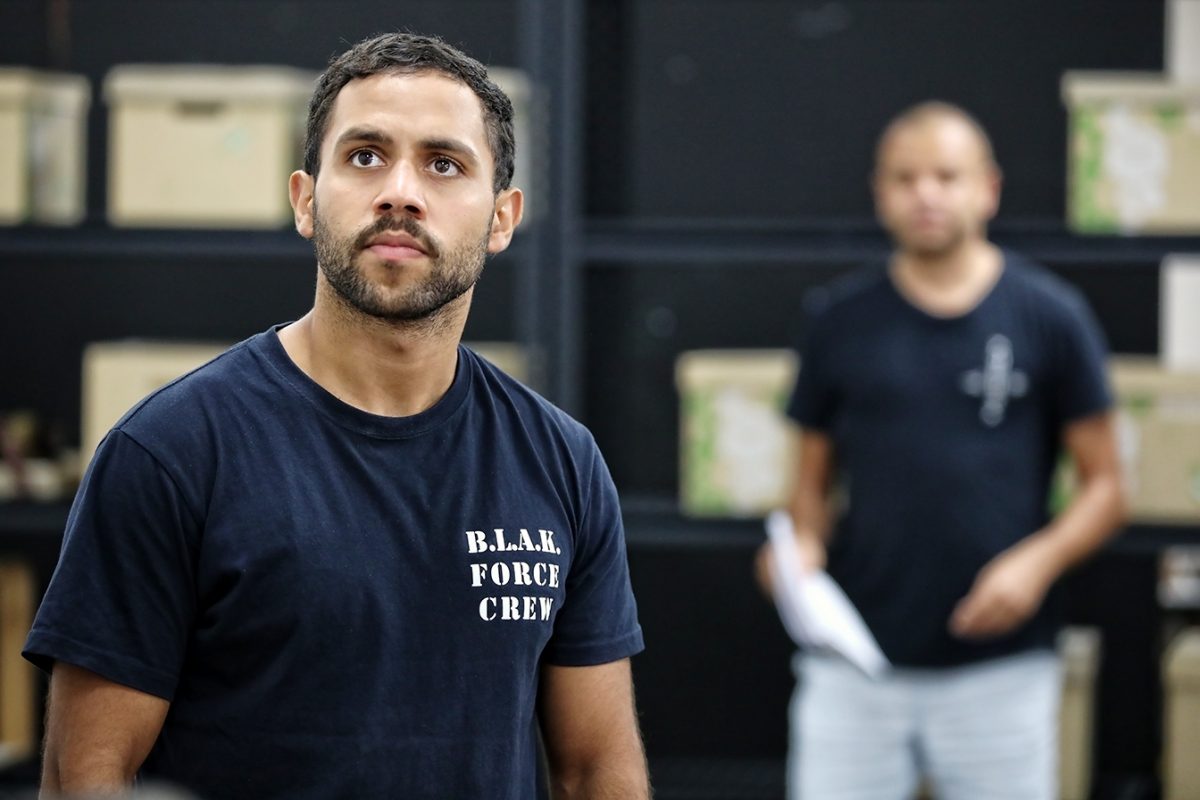 A man wearing a B.L.A.K. FORCE CREW T-shirt stands in focus indoors, looking slightly to the side, with shelves and boxes behind him and another person blurred in the background.