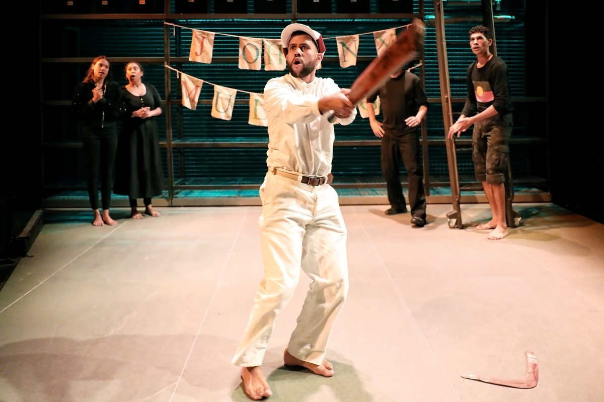 A barefoot man in white clothes and hat swings a bat theatrically on stage, whilst four people watch in the background. A string of papers hangs behind them, suggesting a theatrical or performance setting.