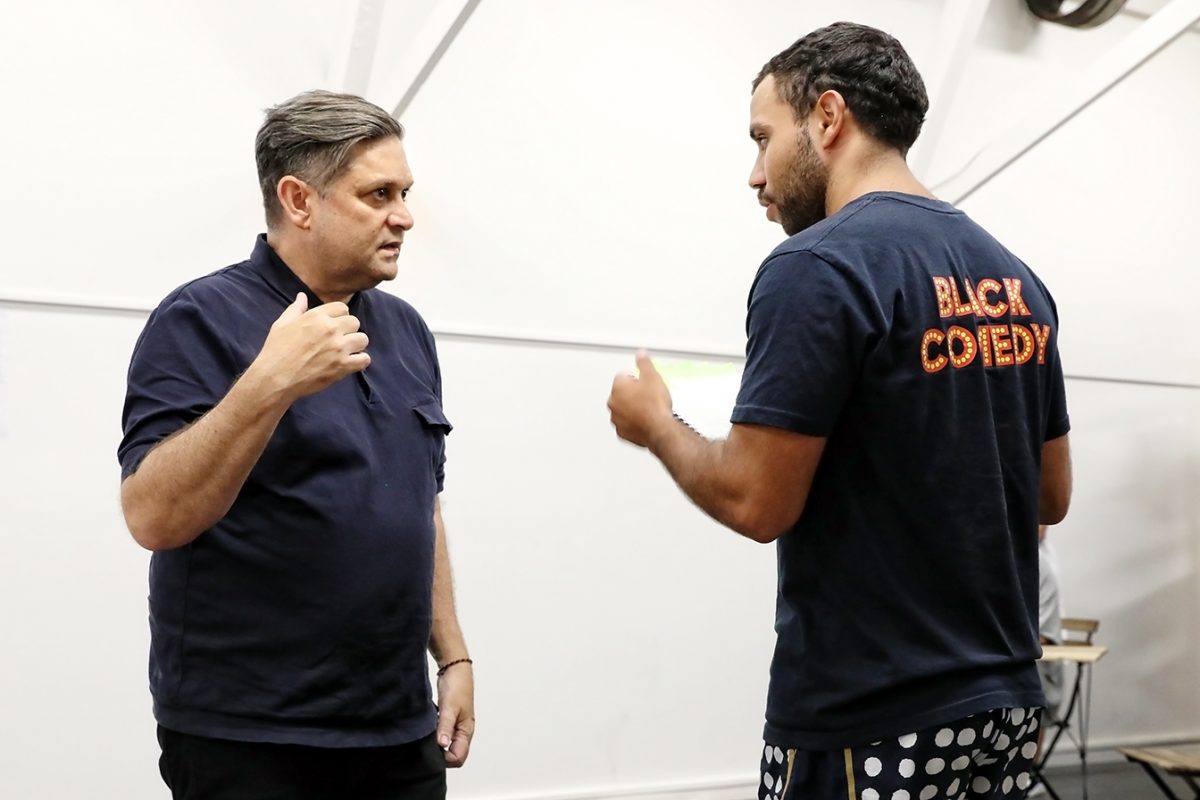 Two men are standing indoors having a conversation. One gestures with his hand, while the other, wearing a shirt that says BLACK COMEDY on the back, listens attentively. The background is minimal and white.
