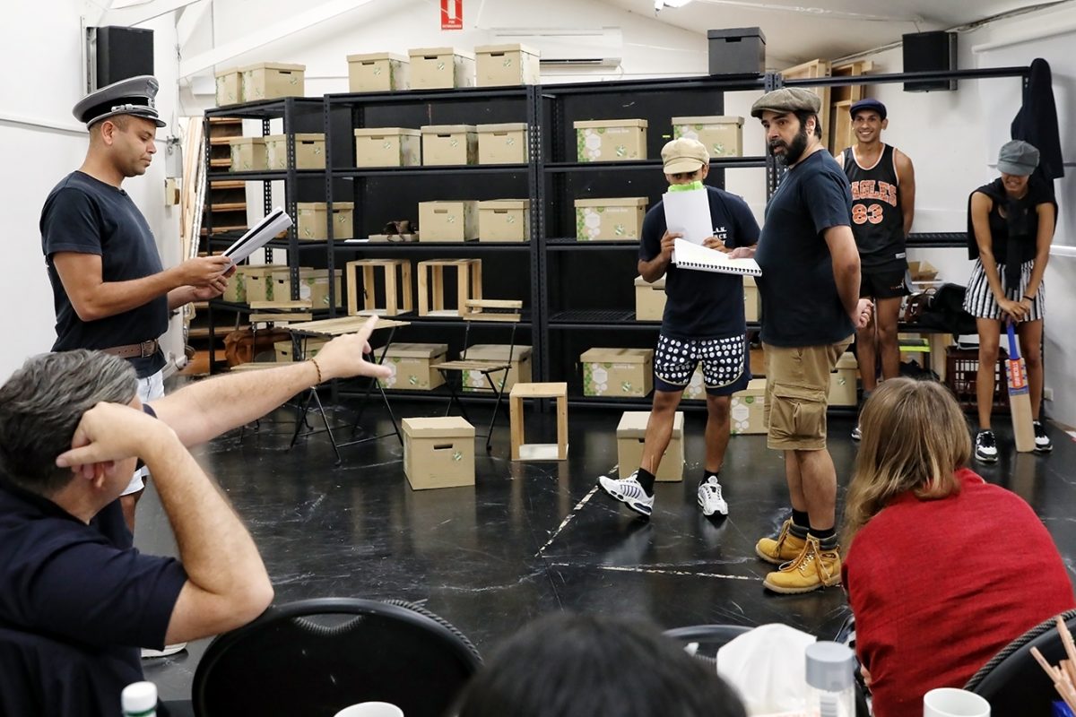 A group of people participate in a script reading or rehearsal in a room lined with shelves of cardboard boxes. Some stand and act, whilst others sit at a table, observing and gesturing towards the actors.