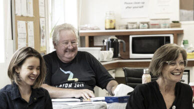 Three adults sitting and smiling in a cosy room with a kitchen area in the background. A man sits at a table with papers and tissues, while two women sit in front, laughing. A sign on the wall reads, “Please tidy up after yourself.”.