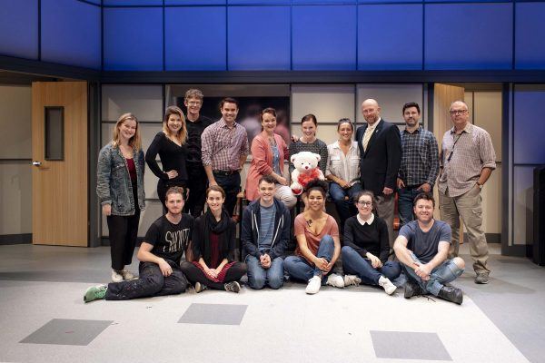 A group of sixteen people pose together indoors, with some standing and others sitting cross-legged on the floor. There's a large teddy bear in the middle, and they're in a modern room with blue lighting and wooden doors.