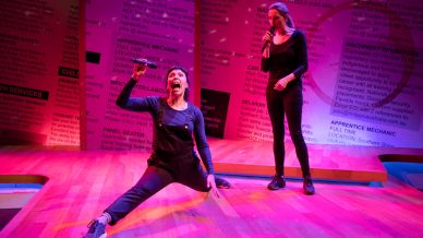 Two performers on stage: one person is doing the splits and singing passionately into a microphone, whilst the other stands watching with a microphone. The background features large, colourful job listing texts.