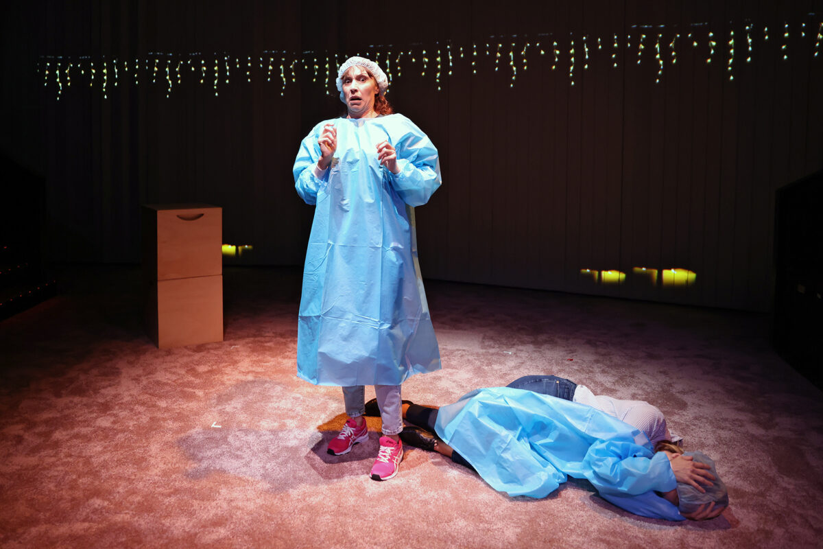 A person in a blue medical gown and hair cover stands on stage, looking concerned, whilst another person lies curled up on the floor behind them under stage lights with fairy lights in the background.