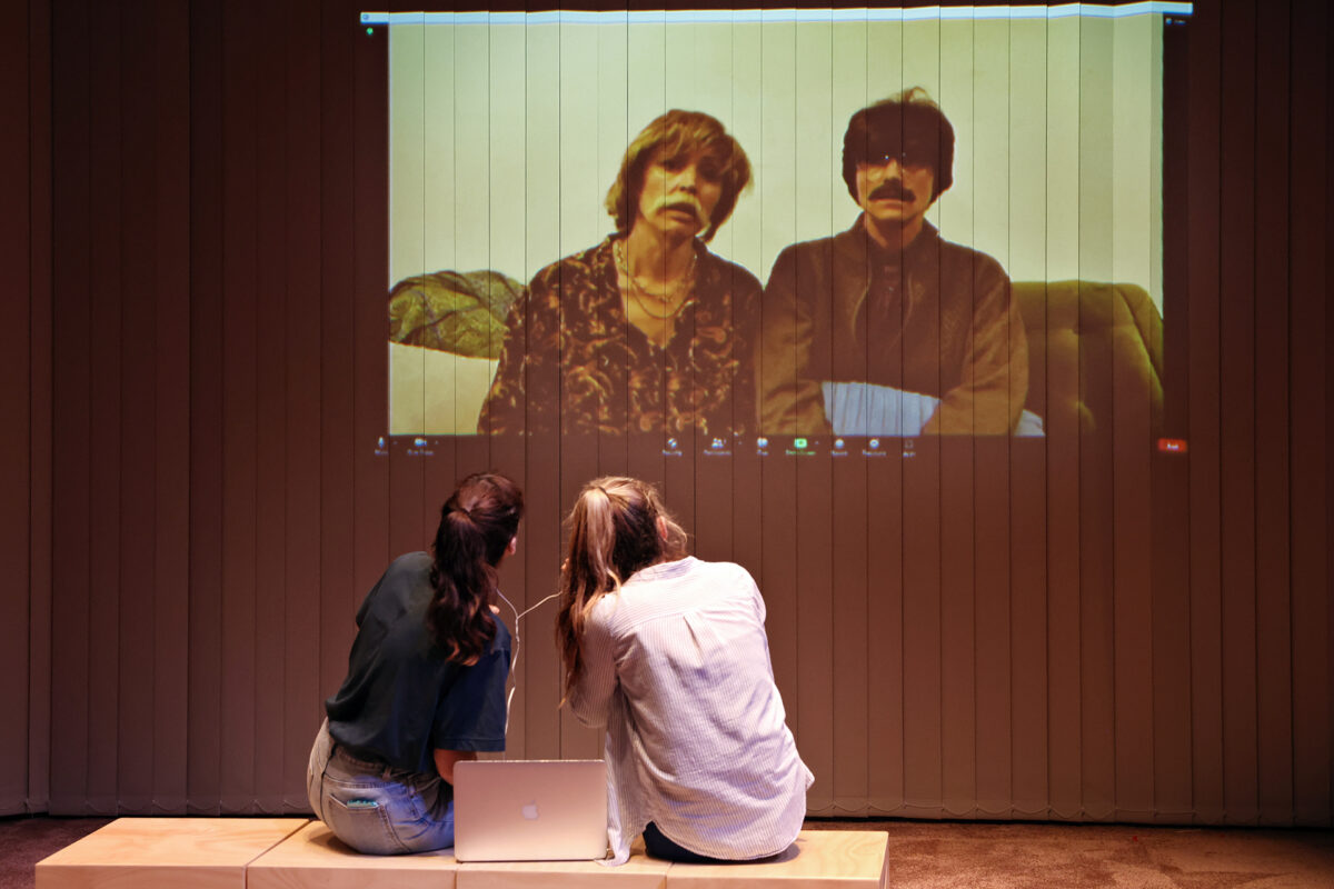 Two people sit on a bench with a laptop, watching a video call projected on a wall showing two other people seated together, both wearing dark sunglasses and patterned clothing.