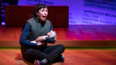 A woman in a green waistcoat and black trousers sits cross-legged on a wooden floor, holding a baby doll and looking up with a surprised expression. A background wall displays large, blurred text and job adverts.
