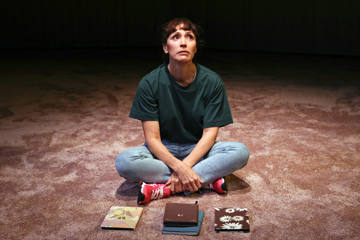 A woman in a green shirt and jeans sits cross-legged on a carpeted floor, looking upwards. In front of her are three closed notebooks with different designs. The lighting is dim and focused on her.