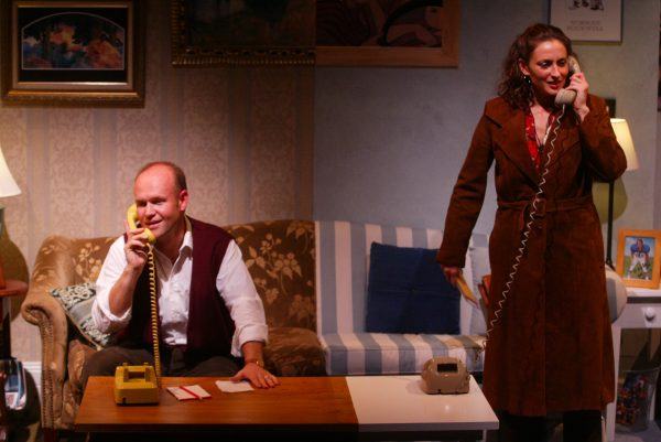 A man and a woman speak on separate corded telephones in a warmly lit, vintage-style sitting room with patterned furniture and framed pictures on the walls. The man sits at a table, while the woman stands nearby, smiling.