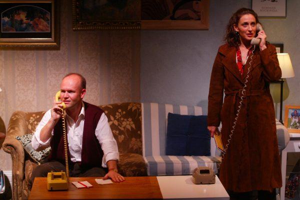 A man sits on a sofa and talks on a yellow rotary phone while a woman in a brown coat stands nearby, also speaking on a beige rotary phone. The setting appears to be a warmly lit lounge.