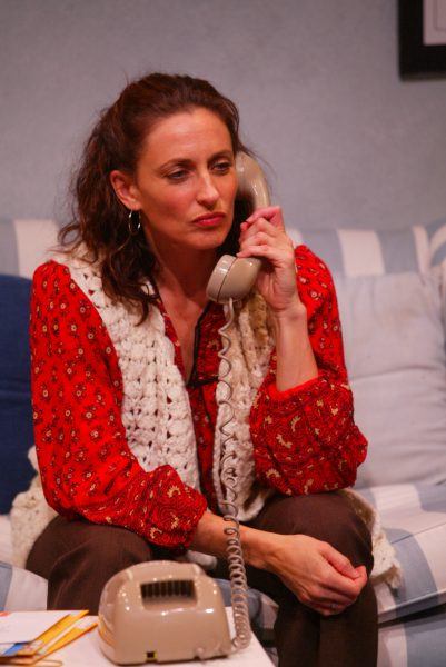 A woman with wavy brown hair, wearing a red patterned blouse and crocheted waistcoat, sits on a striped sofa holding a beige rotary telephone to her ear, appearing thoughtful or concerned.