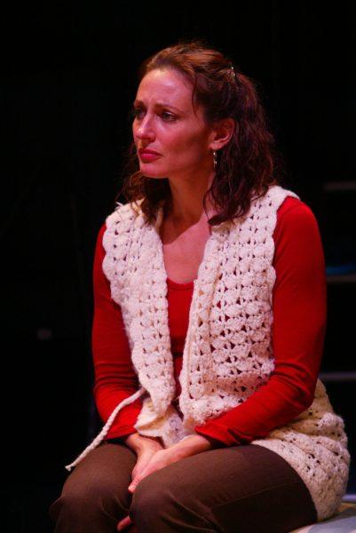 A woman with curly brown hair wearing a red long-sleeved shirt and a white crocheted waistcoat sits with her hands clasped, looking slightly to the side with a serious expression against a dark background.