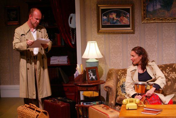 A man in a trench coat reads a paper while a woman in a similar coat sits on a sofa with a drum. They are surrounded by vintage suitcases in a warmly lit, traditionally decorated sitting room.