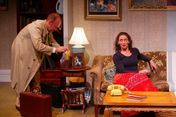 A man in a beige trench coat looks through files at a desk, while a woman with a red patterned skirt sits on a sofa, glancing at him. A yellow telephone and books are on the coffee table in front of her.
