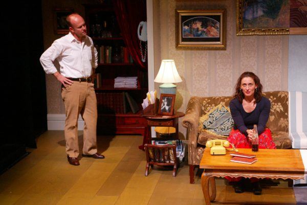 A man stands with hands on hips, looking at a seated woman on a patterned sofa. The room has a lit table lamp, books, a yellow rotary telephone, and framed art on the wall. The woman looks thoughtful at a coffee table.