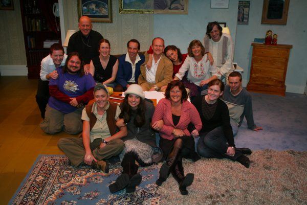 A group of thirteen people, smiling and sitting closely together on a decorative rug and wooden floor in a cosy, warmly lit room with framed art and shelves in the background.