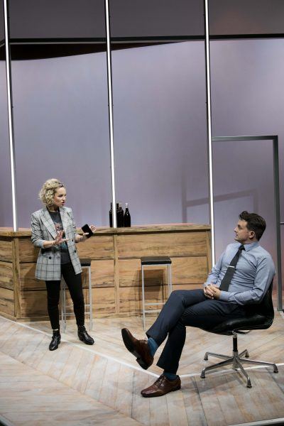 A woman in a checked blazer stands and speaks to a seated man in a suit and tie. They are on a modern stage set with wooden panels, bar stools, and wine bottles in the background.