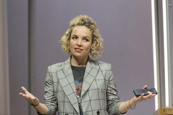 A woman with curly blonde hair wearing a grey checked blazer and holding a mobile phone shrugs her shoulders with an uncertain or confused expression.