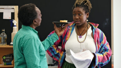 A man in a green shirt holds a plate with a small piece of food near a woman in a white top and colourful jacket, who looks unimpressed while holding papers and a glass. They are in a room with shelves and a black wall.