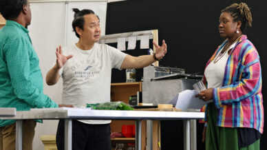 Three people stand in a kitchen or classroom setting. One man speaks animatedly, gesturing with open hands, whilst another man and a woman listen attentively. Papers and kitchen equipment are on the table in front of them.
