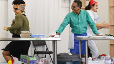 Three people in a kitchen setting smile and dance while standing beside metal tables with food containers. One wears a headscarf, another has a towel on their shoulder, and the third has a blue apron. The mood is lively and fun.