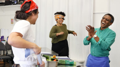 Three people in a bright kitchen laugh and chat together. One wears a red bandana, another a colourful headwrap, and the third a blue apron. Fresh vegetables and kitchen tools are on the counter in front of them.