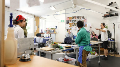 A group of people are gathered in a bright, cluttered room, listening to a person standing and gesturing at the front. Various items and supplies are on tables, and a noticeboard with papers is visible on the back wall.