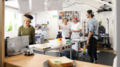 Four people stand and smile in a bright, casual kitchen or café. A woman at the counter grins, while three others, dressed in kitchen attire, interact behind her. A sandwich sits on the counter in the foreground.