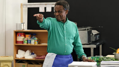 A man in a green shirt and blue apron stands behind a counter, smiling and pointing forward. Behind him are shelves with food items, kitchen roll, and kitchen equipment.